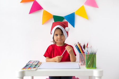 A child in a Santa hat and red shirt sitting at a table with art supplies, preparing to draw, symbolizing creativity and festive joyの写真素材