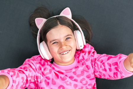 Smiling child girl 8-9 years in cat ear headphones lying on trampoline in pink leopard sweatshirt, looking up with joy, concept of childhood fun and technologyの写真素材