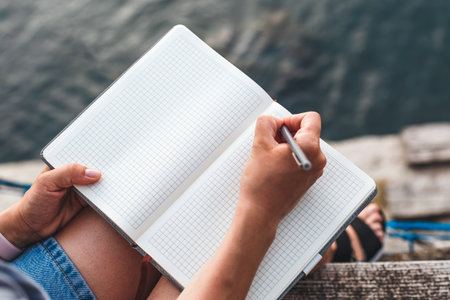 Woman writing with pen in blank notebook while sitting on wooden pier over calm water, wearing blue...