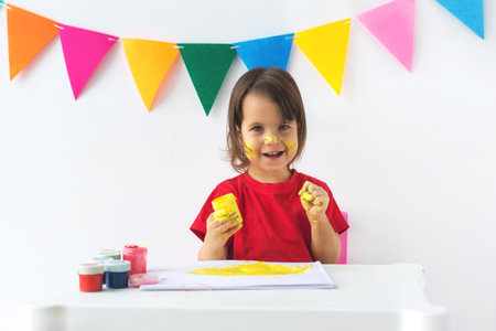 A cheerful little girl 2-3 year in a red t-shirt with yellow paint on her face and hands smiles while holding a paint jar. A festive background with colorful flags enhances the joyful atmosphereの写真素材