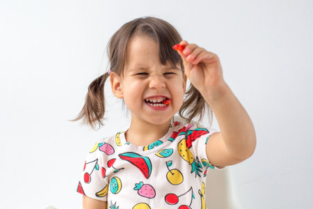 Little girl with pigtails sitting in a high chair, joyfully holding up a piece of tomato while making a playful grimace. enjoying her snack.の写真素材