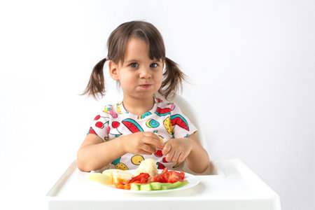 Little girl with pigtails sitting in a high chair, eating fresh vegetables and making a funny face. Healthy eating habits, childhood nutrition, and balanced diet. Concept of childhood and healthの写真素材