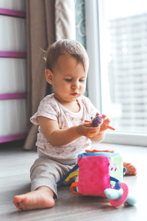 Baby girl in floral bodysuit playing with soft cube toy near window on wooden floor, looking at...