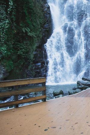 vertical photo cascading waterfall behind a wooden deck with a railing in the mountainous region of Adjara, Georgia. lush greenery adds to the tranquil atmosphere. Concept of nature and tranquilityの写真素材