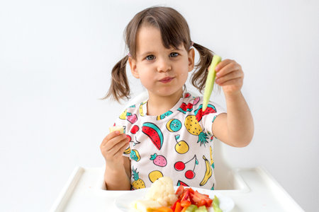 Little girl with pigtails sitting in a high chair, eating fresh vegetables and holding a cucumber stick while looking at it. Healthy eating habits for children.の写真素材