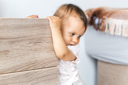 Baby holding onto wooden furniture while standing indoors, looking thoughtful and curious, symbol of...