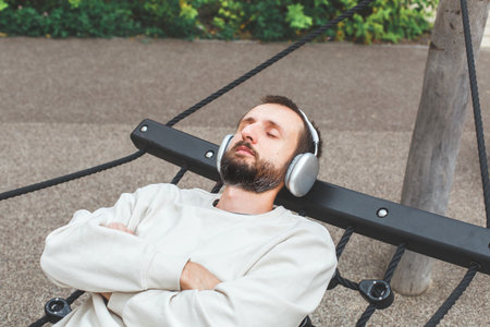 Young man with beard relaxing outdoors in hammock, wearing wireless headphones and enjoying music with eyes closed. Concept of relaxation, mindfulness, stress relief and mental well-being.の写真素材
