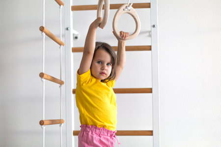 Little girl about four years old hanging on wooden gymnastic rings at home, developing coordination, strength, and confidence through active play and early physical educationの写真素材
