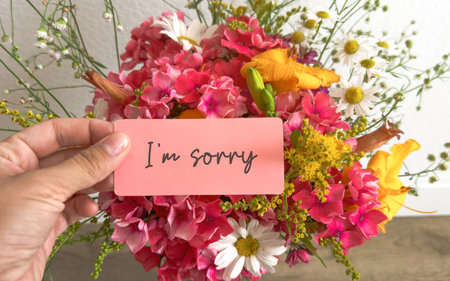 Hand holding a pink card with words text I m sorry against a vibrant bouquet of mixed flowers...