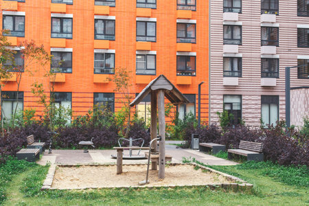 Small playground with sandpit, wooden play equipment, benches and greenery between colorful modern apartment buildings, symbolizing family leisure, childhood, safety and urban comfortの写真素材