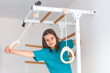 Smiling girl in turquoise t-shirt playing on indoor gymnastic rings at home, developing strength and...
