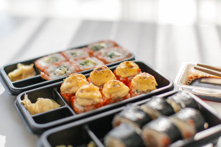 Assorted sushi rolls with baked toppings in plastic delivery boxes served with soy sauce, ginger and chopsticks, concept of Japanese cuisine, food delivery and modern dining at homeの写真素材