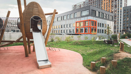 Modern urban playground with wooden rocket-shaped slide and climbing structures surrounded by green grass, located in residential courtyard near contemporary apartment buildings and schoolの写真素材