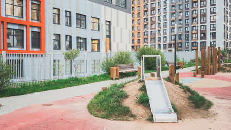 Modern courtyard playground with small metal slide built into grassy hill, wooden benches and play elements surrounded by residential buildings with colorful facades and urban landscapingの写真素材