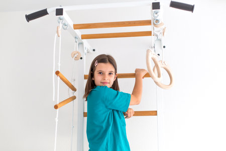 Smiling child girl with long brown hair turquoise T-shirt climbing a wooden gym ladder indoors, with gymnastic rings and ropes nearby. Concept of childhood, fitness, health, activity, home exerciseの写真素材