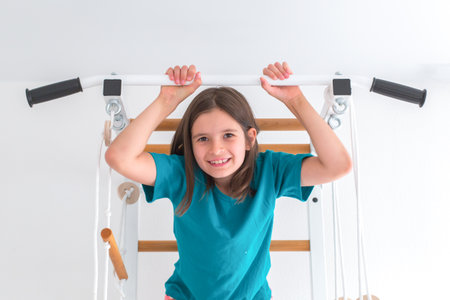 Smiling girl with long brown hair in a turquoise T-shirt hanging on a pull-up bar indoors, looking...
