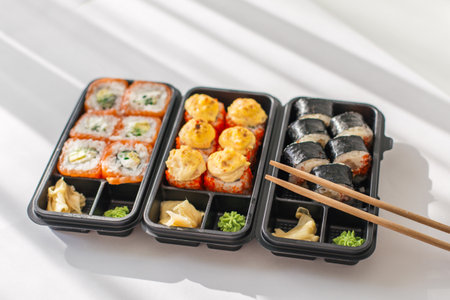 Assorted sushi rolls in black plastic containers with ginger, wasabi, and wooden chopsticks on a...
