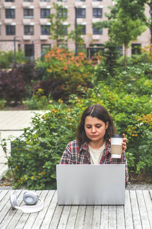 A vertical portrait of young Caucasian female freelancer working or studying in front of a laptop and talking on the phone in the courtyard of a residential complex on a warm dayの写真素材