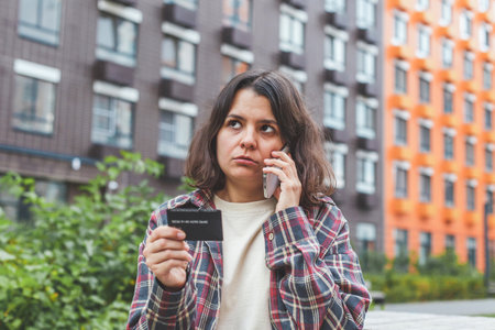 Woman standing outdoors, talking on the phone and holding a bank card, looking worried and frustrated. Concept of financial problems, card blocking, fraud alert, or customer service communicationの写真素材