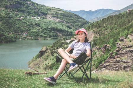 Girl in pink cap sitting on camping chair on grassy hill overlooking green mountain lake, relaxing and enjoying summer day in nature, symbolizing childhood freedom, calmness and outdoor recreationの写真素材