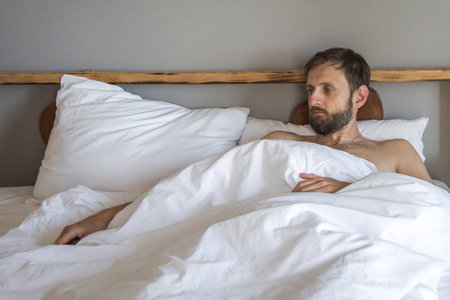 bearded man lying in bed under white blankets, looking thoughtful and tired, symbolizing morning laziness, insomnia, low energy, or emotional exhaustion in a cozy bedroom interiorの写真素材