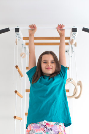 Cheerful girl with brown hair and turquoise T-shirt hanging on a pull-up bar indoors, smiling...