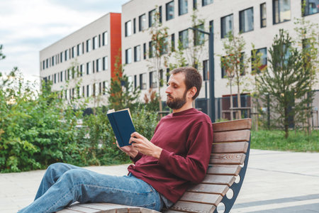 Young man in burgundy sweatshirt sitting on modern wooden bench outdoors, reading book near contemporary buildings with greenery, concept of education, relaxation modern urban infrastructureの写真素材