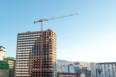 Modern high-rise building under construction with red tower crane against clear blue sky, surrounded by other contemporary buildings mountains in distance, concept of urban development architectureの写真素材