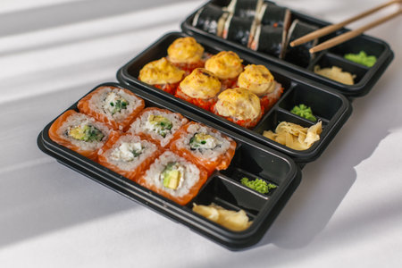 Assorted sushi rolls in black plastic containers with wasabi, pickled ginger and chopsticks on a white table, concept of Japanese cuisine, takeaway food and healthy eatingの写真素材