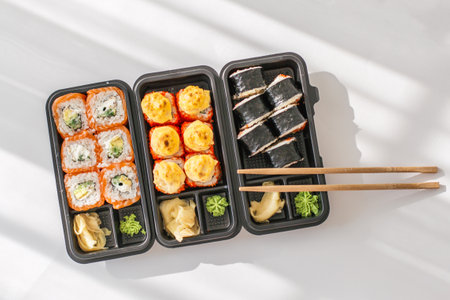 Assorted sushi rolls in black takeout boxes with chopsticks, wasabi and ginger on white background, showcasing fresh Japanese cuisine perfect for restaurant menus, delivery ads and food blogsの写真素材