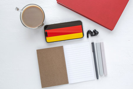 Flat lay with smartphone showing German flag, notebook, pens, coffee cup and earbuds on white desk, symbolizing learning German language, education, culture and international communicationの写真素材