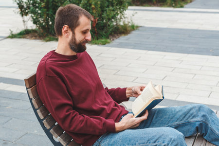 Young man in burgundy sweatshirt and jeans sitting on wooden bench outdoors reading a book, symbolizing relaxation, knowledge, learning, and peaceful urban lifestyleの写真素材