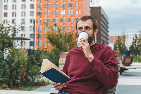 Man beard sitting on a wooden bench outdoors near modern buildings, wearing a burgundy sweatshirt and jeans, holding a book and drinking coffee, concept of urban lifestyle, relaxation and educationの写真素材