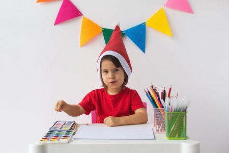 A young child in a Santa hat and red shirt sits at a table with painting supplies, ready to create holiday artwork, symbolizing Christmas creativity and festive joyの写真素材