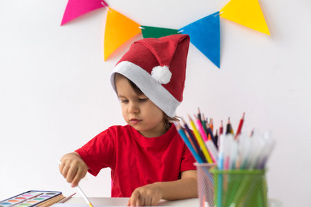 A young child wearing a Santa hat and red shirt painting with watercolors at a decorated table, symbolizing holiday creativity and festive joyの写真素材