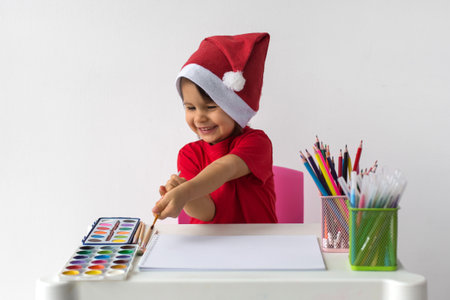A cheerful little girl wearing a Santa hat and red t-shirt is sitting at a table with art supplies, ready to paint with watercolors. She is excited and happy, embracing the holiday spirit. Concept of Christmas creativityの写真素材