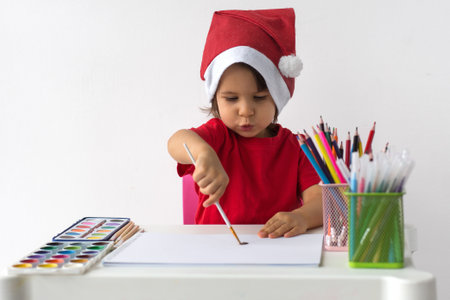 A little girl in a Santa hat and red t-shirt is painting with watercolors at a table filled with art supplies. She is focused on her creative work, enjoying the holiday spirit. Concept of Christmas creativityの写真素材