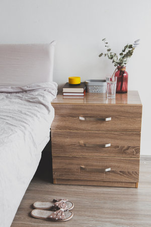 Cozy bedroom interior with wooden bedside table, stack of books, red vase with eucalyptus branches, glass of water yellow candle, polka dot slippers on the floor concept of home comfort, relaxationの写真素材