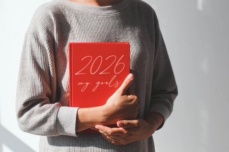 Person in a gray sweater holding a red planner with the inscription 2026 my goals against a bright background, concept of motivation, planning and setting personal objectives for the future yearの写真素材
