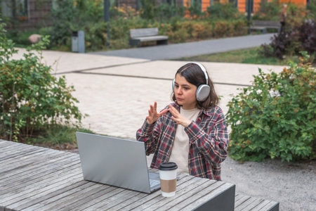 Young woman in a plaid shirt and large headphones sitting outdoors with a laptop, gesturing during an online meeting or video call, concept of remote work, communication and digital lifestyleの写真素材