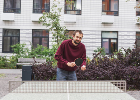 Man in a burgundy sweatshirt and jeans preparing to play table tennis outdoors in a modern...