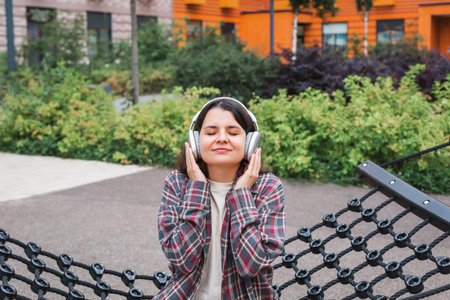 Young woman in plaid shirt headphones sitting outdoors in a modern residential courtyard, closing her eyes and smiling enjoying music. Concept of relaxation, comfort, happiness, modern urban lifestyleの写真素材
