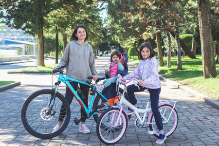 Smiling woman with two children on bicycles in sunny park alley, one child sitting in bike seat....