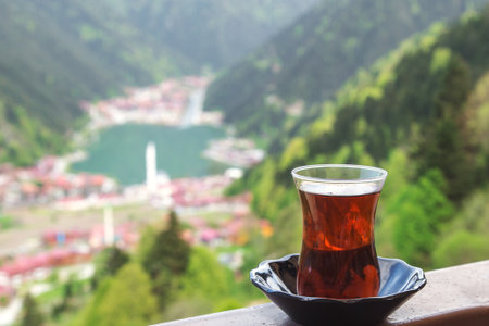 Glass of hot black tea on a balcony overlooking a picturesque mountain valley with a lake and village below. Concept of travel, relaxation, nature, and traditional tea cultureの写真素材