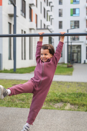 Little girl in a burgundy tracksuit hanging on a horizontal bar outdoors, smiling and lifting one...