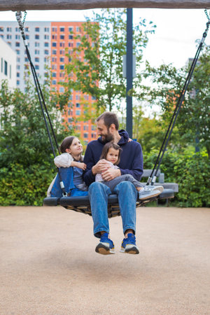 Father with two daughters sitting together on a large swing in a modern playground of a residential area, smiling and spending quality time outdoors, concept of family love, bonding and happy parenthoodの写真素材
