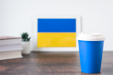 National flag of Ukraine on the tablet, textbooks, a blue cup of hot drink coffee or tea on the table, the concept of learning Ukrainian languageの写真素材