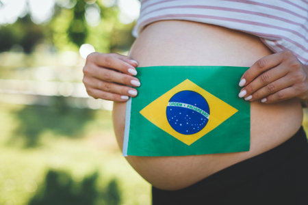 Pregnant woman holding the Brazilian flag on her bare belly, symbolizing national pride and motherhood. The outdoor setting adds warmth and natural beautyの写真素材