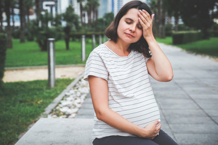 a sad pregnant woman sits alone on a bench in the park in summer holds on to her head and belly, discomfort and pain during pregnancyの写真素材