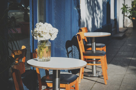 A cozy outdoor cafe with wooden chairs and round tables, featuring a vase of fresh white flowers in sunlight. blue building facade adds charm and warmth to the inviting atmosphere.の写真素材